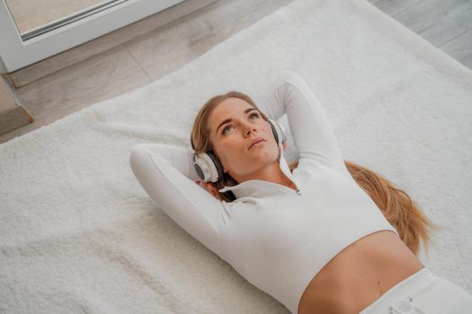 Top view portrait of relaxed woman listening to music with headphones lying on carpet at home. She is dressed in a White tracksuit