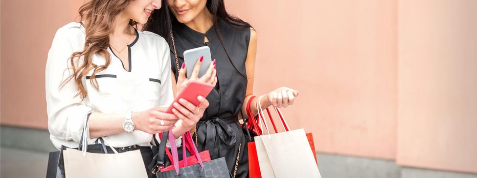 Two beautiful caucasian young women watching at smart phone on new seasonal offers with bags during shopping near wall of shopping mall