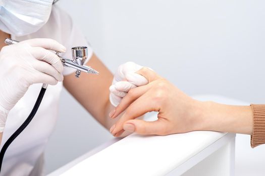 Young woman receiving manicure by airbrush in nail salon. Procedure for spraying paint on the nails