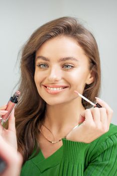 Beautiful young caucasian smiling woman applying gloss to the lips looking in the mirror