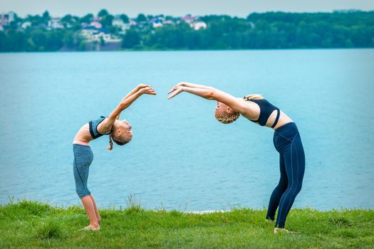 Mother and daughter doing gym exercises on the grass at the shore of the lake