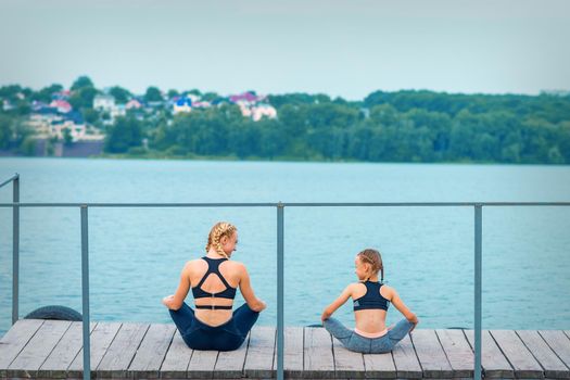 Mother and daughter doing gym exercises on the grass at the pier of the river