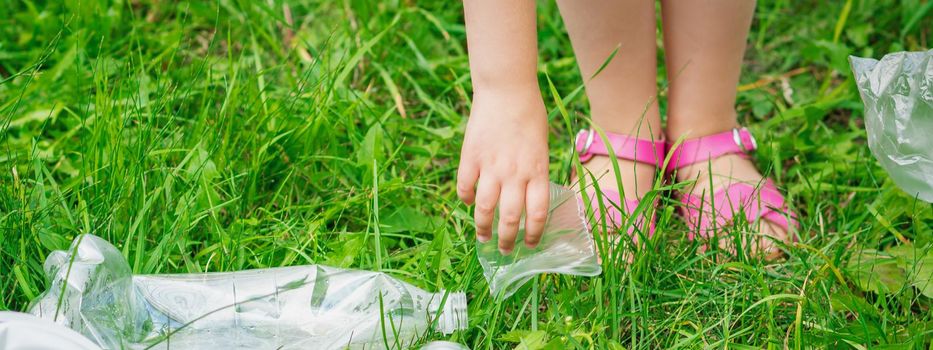 Hand of child cleans green grass from plastic trash in the park