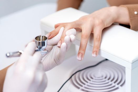 Young woman receiving manicure by airbrush in nail salon. Procedure for spraying paint on the nails