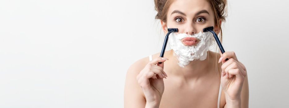 Beautiful young caucasian woman shaving her face by two razors on white background. Pretty woman with shaving foam on her face