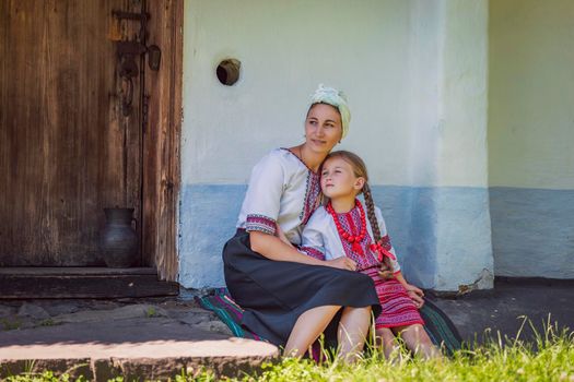 mother and daughter in Ukrainian national costumes are sitting near an old house