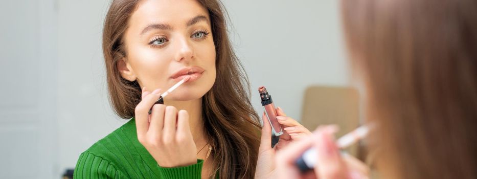 Beautiful young caucasian woman applying gloss to the lips looking in the mirror. Out of focus