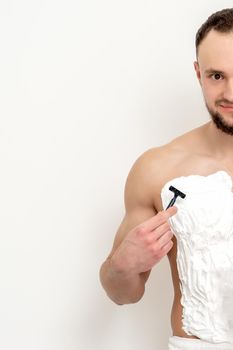 Young caucasian man with beard holds razor shaves his chest with white shaving foam on white background. Man shaving his torso