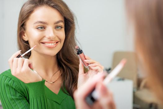 Beautiful young caucasian smiling woman applying gloss to the lips looking in the mirror