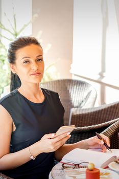 Portrait of young business woman makes notes in notebook from smartphone working at coffee break in cafe