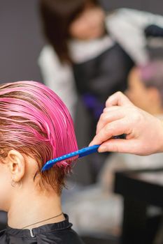 Young woman receiving hair treatment after pink coloring by hand of male hairdresser in hair salon