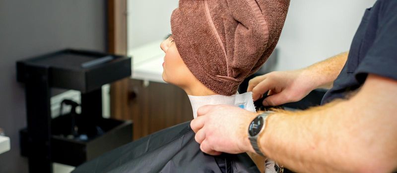 Hairdresser puts on black cape to female customer with towel on her head in hair salon