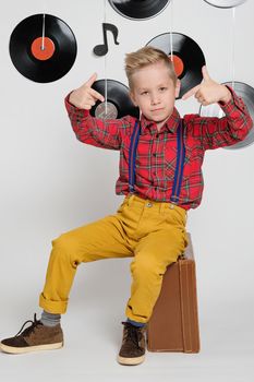 Retro disco 60s, 70s, 80s concept, funny boy wearing checked shirt, yellow trousers and stylish haircut sitting on a suitcase, background with music plate