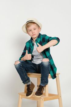 Portrait of cute stylish blond boy kid 7 years old in checked shirt and jeans sitting on a ladder, with arms spread in