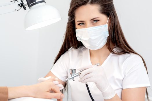 Portrait of manicure master uses airbrush wearing mask in nail salon