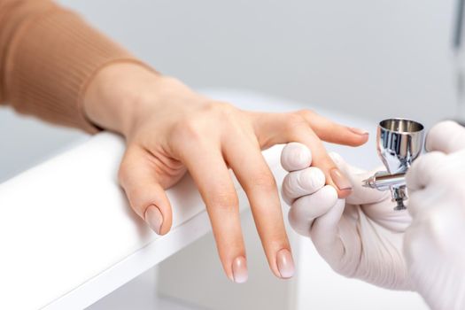 Young woman receiving manicure by airbrush in nail salon. Procedure for spraying paint on the nails