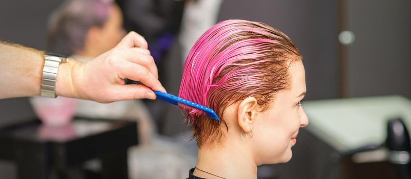 Young woman receiving hair treatment after pink coloring by hand of male hairdresser in hair salon