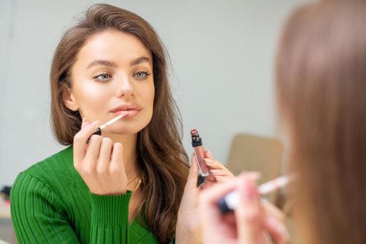 Beautiful young caucasian woman applying gloss to the lips looking in the mirror