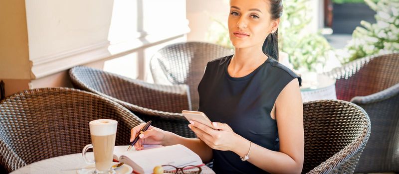 Portrait of young business woman makes notes in notebook from smartphone working at coffee break in cafe