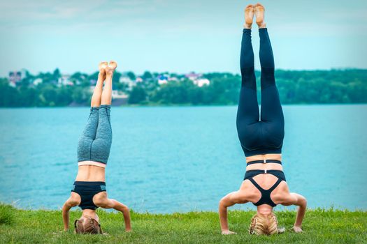 Mother and daughter doing gym exercises on the grass at the shore of the lake. Woman and girl stands on head