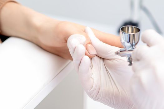 Young woman receiving manicure by airbrush in nail salon. Procedure for spraying paint on the nails