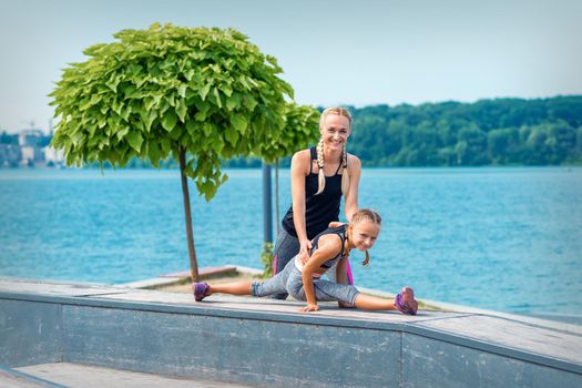 Mother and daughter doing gym exercises on the grass at the pier of the river