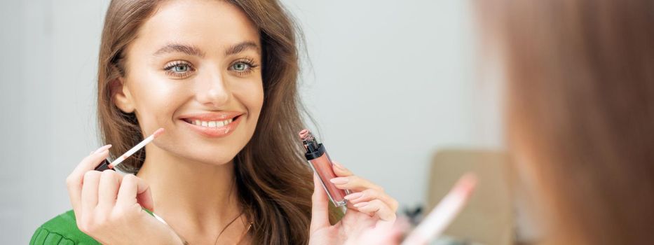 Beautiful young caucasian smiling woman applying gloss to the lips looking in the mirror