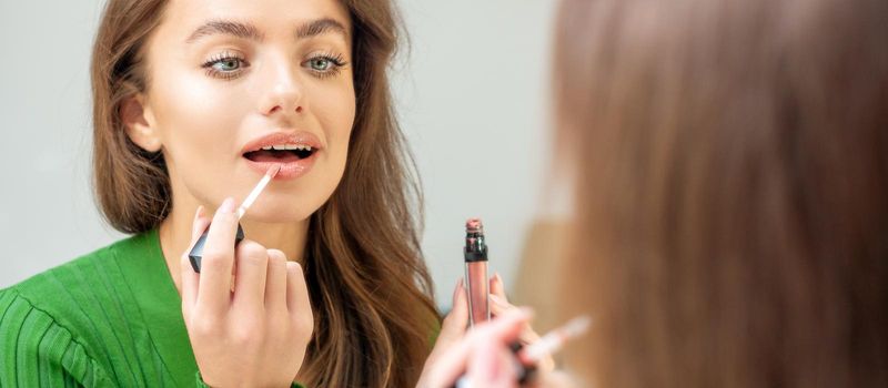 Beautiful young caucasian woman applying gloss to the lips looking in the mirror