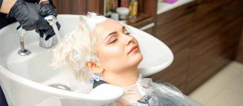 Hairdresser prepare to washes off the white dye from hair of young caucasian woman in sink at beauty salon