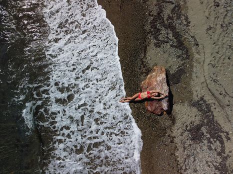 Young sensual blonde woman sitting on the rock near water at sea and enjoys the sea waves on background of two volcanic rocks, like in Iceland. Dreams holidays and weekend vacation in summer time