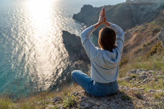 Woman tourist enjoying the sunset over the sea mountain landscape. Sits outdoors on a rock above the sea. She is wearing jeans and a blue hoodie. Healthy lifestyle, harmony and meditation.