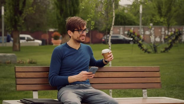 Handsome young man browsing smartphone social network app smiles texting a message while drink coffee. Student man relaxing in a park.