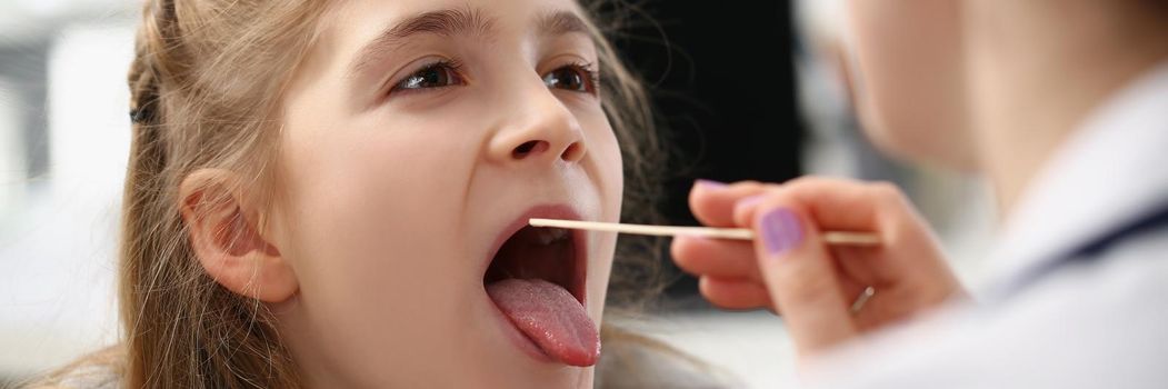 Portrait of little girl having her throat examined with special spoon at woman doctor in hospital. Child open mouth for investigation. Healthcare concept