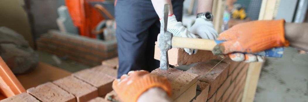 Close-up of qualified worker hit on red brick with hammer tool for better fixation. Group of workers building new house project. Construction site concept