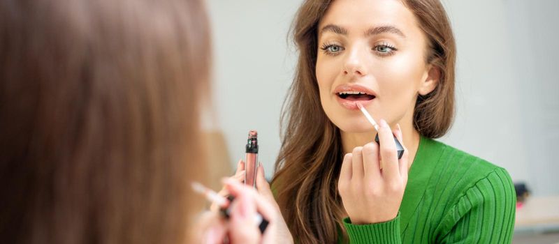 Beautiful young caucasian woman applying gloss to the lips looking in the mirror