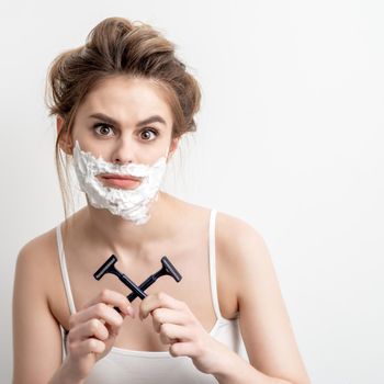 Beautiful young caucasian woman with shaving foam on her face and two razors in her hands on white background