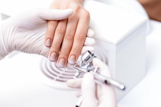 Young woman receiving manicure by airbrush in nail salon. Procedure for spraying paint on the nails