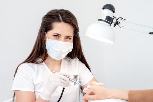 Portrait of manicure master uses airbrush wearing mask in nail salon