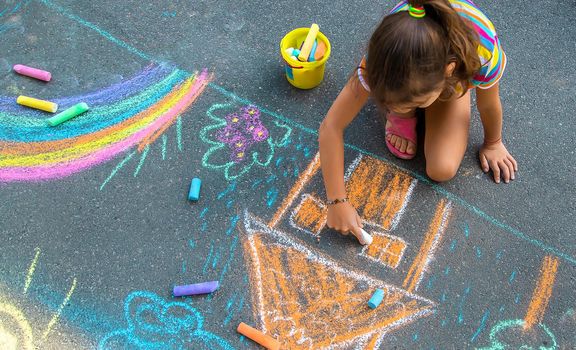 The child draws a house and a rainbow on the asphalt with chalk. Selective focus. Kids.