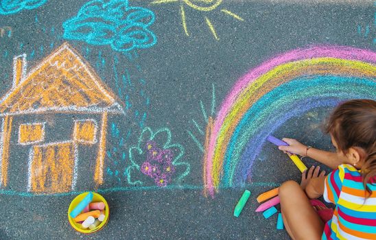 The child draws a house and a rainbow on the asphalt with chalk. Selective focus. Kids.