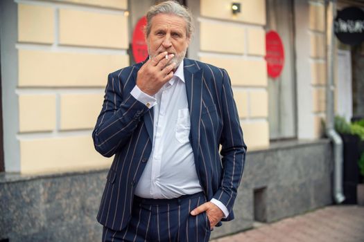 an elderly man smokes in the street with one hand in his pants pocket