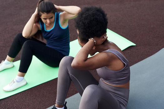 Cheerful young diverse female friends doing ab crunches on stadium in the park - friends together doing exercise outdoors.