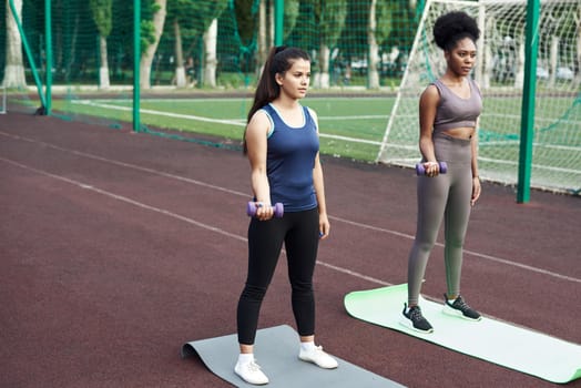 Happy women exercising with dumbbells outdoors. Fitness women doing a workout in the stadium and working out with small dumbbells.