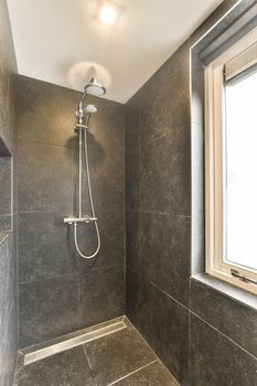 Modern black shower column in expensive open bathroom with gray tiled wall and window