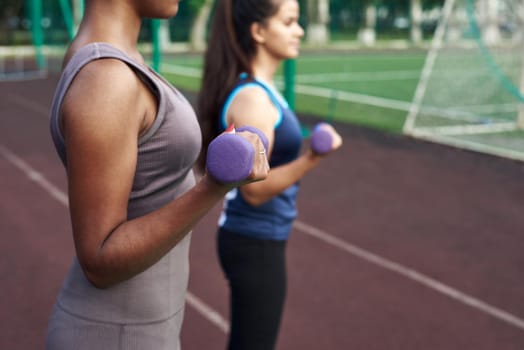 Young diverse women pump arm muscles with dumbbells in the outdoor stadium. Close up.