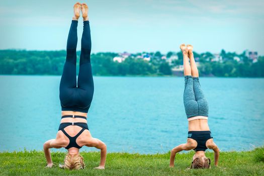 Mother and daughter doing gym exercises on the grass at the shore of the lake. Woman and girl stands on head
