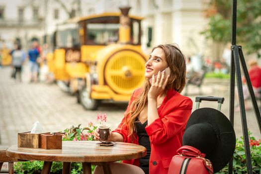 Beautiful happy young caucasian woman talking by cell phone sitting in outdoors cafe at european city