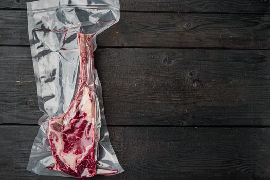 Dry aged raw tomahawk beef steak in plastic airtight pack set, on black wooden table background, top view flat lay, with copy space for text