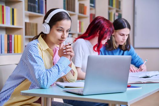 Group of teenage students study in school library classroom. In focus is teenager girl in headphones using laptop. High school, learning, education, knowledge, adolescence, people concept