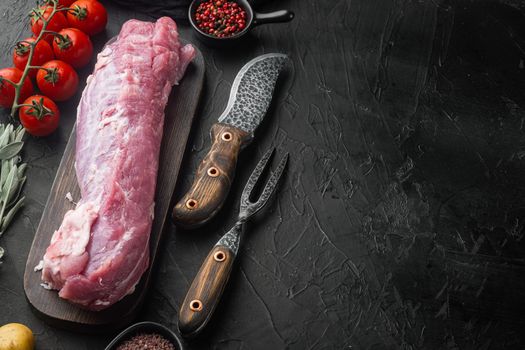 Whole boneless pork fillet loin with ingredients and herbs for grill set, on wooden cutting board, with barbeque knife and meat fork, on black stone background , with copyspace and space for text